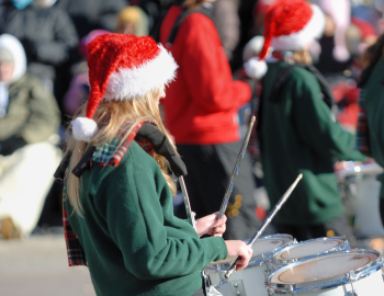 Christmas Parade Person wearing a santa hat and green sweater playing tenor drums in a parade.