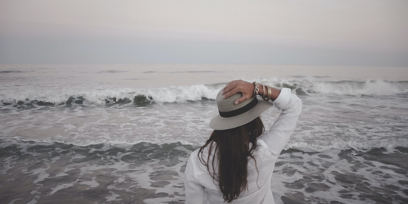 Dress in Layers Woman standing in front of gray ocean with hat and sweater.