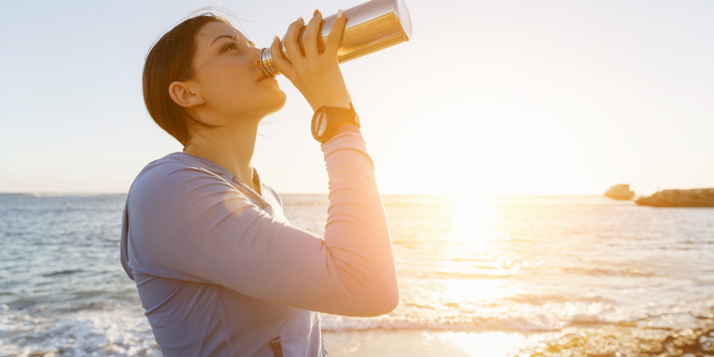 Stay Hydrated Woman drinking out of silver waterbottle at the beach.
