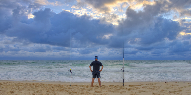 Fishing Man standing on the beach with fishing poles in the sand.