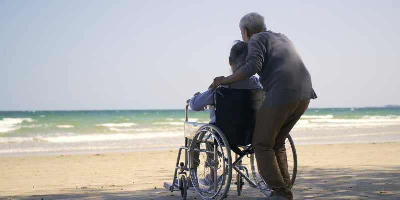 Everyone Deserves an Amazing Outer Banks Vacation! Woman pushing man in wheelchair through the sand towards the ocean.