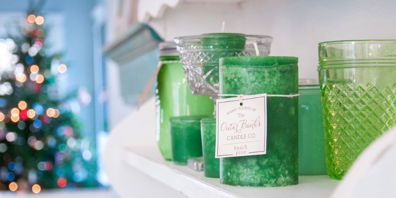 Outer Banks Candle Company Green candles sitting on a shelf with an out-of-focus Christmas tree in background.