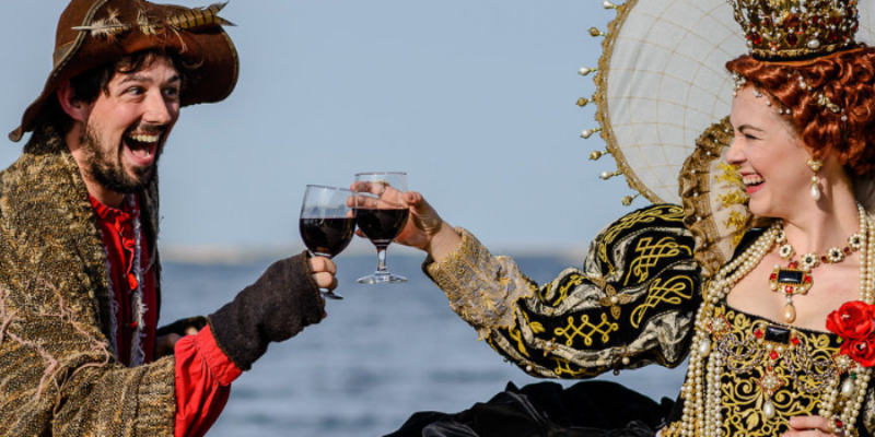 Lost Colony Culinary Festival Image of actress dressed up as Queen Elizabeth and man in a large hat toasting wine glasses.
