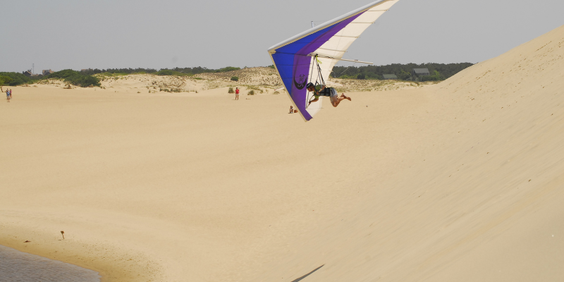 Image of large sand dune; man hang gliding from dune with blue, purple, and white hang glider.