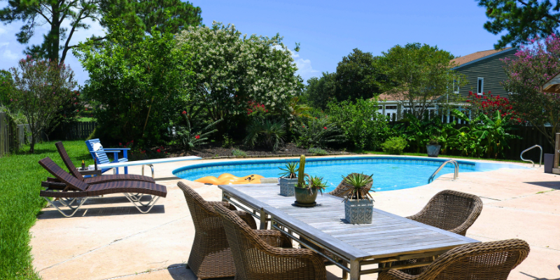 Swimming pool in front of foliage with a dining table and two lounge chairs on the deck
