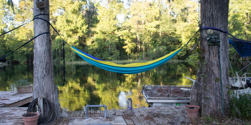 Blue and yellow hammock between two trees in front of a river and treeline.
